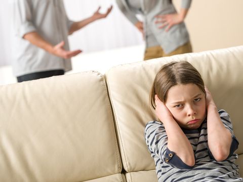 Mädchen sitzt auf Sofa und hält sich die Ohren zu, Mutter und Vater streiten sich im Hintergrund