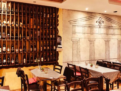 Italienisches Restaurant. Im Vordergrund ein Tische und Stühle in dunklem Holz. Dahinter an der Wand dunkle Weinregale mit Weinflaschen
