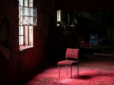 Ein Stuhl steht vor einem Fenster in einer Industrieruine. Alles ist mit rotem Pulver überzogen.