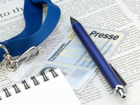 Auf einer Zeitung liegen ein Presseausweis, ein blauer Kugelschreiber und ein Notizblock