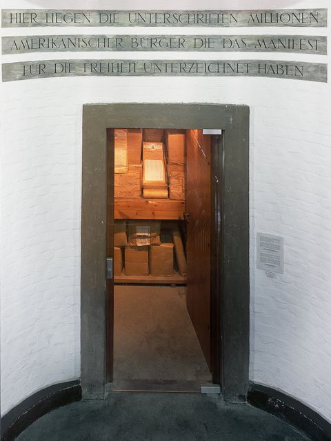 Bildvergrößerung: Offene Tür mit einer Inschrift über dem Eingang, das Innere ist beleuchtet und zeigt in braunes Papier gewickelte Pakete.