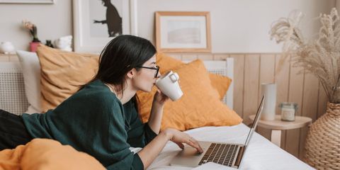 Eine junge Frau liegt auf dem Bett mit einer Tasse und arbeitet am Laptop