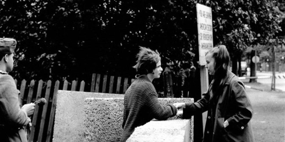 Zwei Mädchen an der noch nicht fertiggestellten Berliner Mauer in Treptow in der Harzer Straße. Sie sprechen miteinander und fassen sich an. Etwas weggedreht steht ein Soldat. Er hat keine Pistole und kein Gewehr in der Hand, sondern eine Blume.