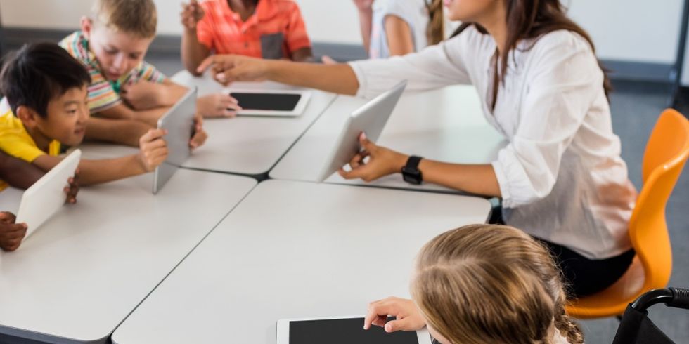 Eine Gruppe von Kindern sitzen mit Tablets in der Hand in einem Klassenraum, ihre Lehrerin zeigt etwas auf ihrem Tablet