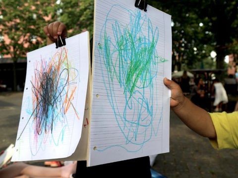 Dotty Tool - der Kunstanhänger draußen unterwegs - Zwei Hände halten bunte Bilder von Kindern auf liniertem Papier in die Kamera