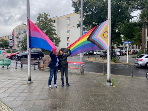 Bildvergrößerung: Diversity- und Queerbeauftragte, Sabine Pöhl, und Bezirksstadträtin Filiz Keküllüoğlu (Bündnis 90 / Die Grünen) halten die beiden Fahnen.