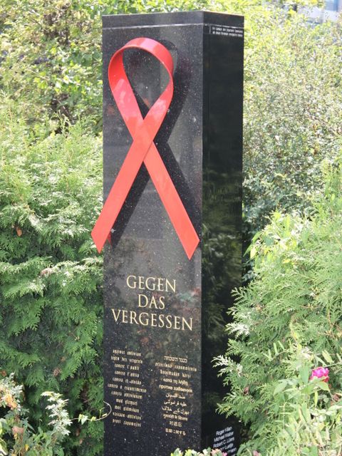 Bildvergrößerung: Eine Stele mit einer großen roten AIDS-Schleife steht zwischen Büschen. Sie trägt die Aufschrift "Gegen das Vergessen".