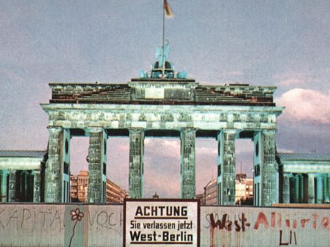 Ein Blick auf die Mauer und das dahinterliegende Brandenburger Tor. Auf einem Schild vor der Mauer steht: Achtung! Sie verlassen jetzt West-Berlin.