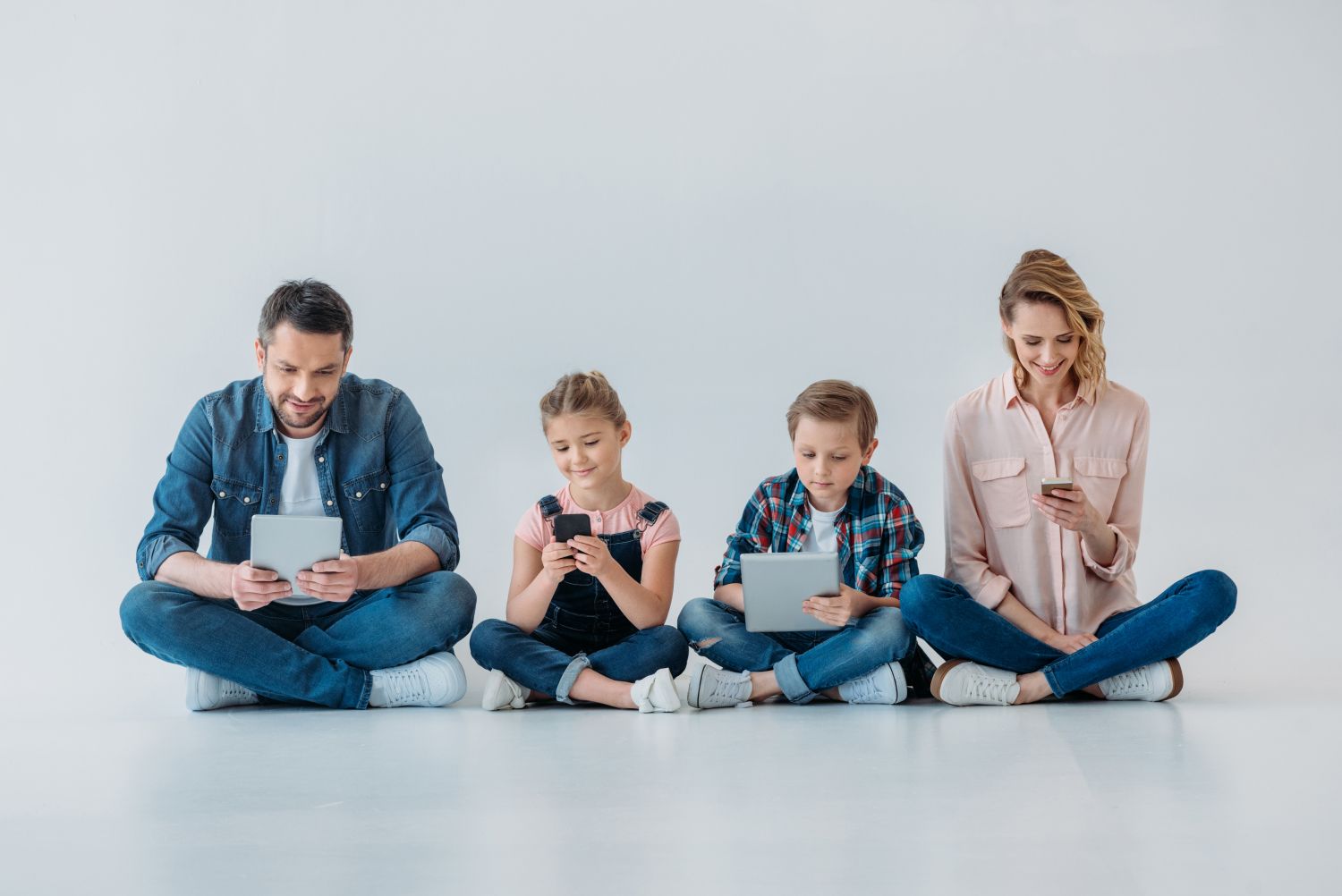 Eltern mit zwei Kindern sitzen auf dem Boden und halten jeweils ein Handy oder ein Tablet