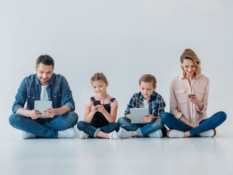 Eltern mit zwei Kindern sitzen auf dem Boden und halten jeweils ein Handy oder ein Tablet