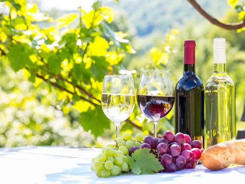 Bildvergrößerung: Ein Tisch im freien auf dem zwei verschiedene Sorten Wein in 2 Flaschen und in 2 Gläsern stehen, davor 2 Sorten Weintrauben und ein Baguette