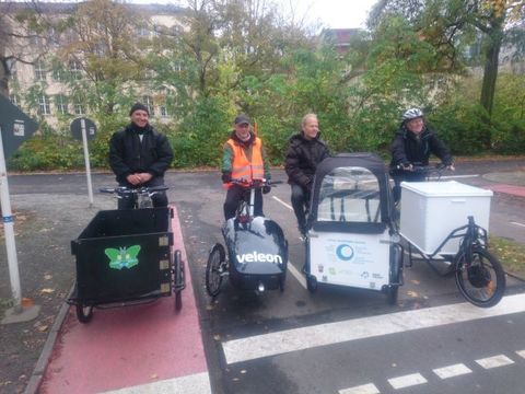 4 verschiedene Lastenfahrräder mit Nutzern auf einer Straße, im Hintergrund Bäume