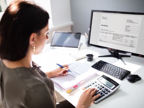 Eine Frau an ihrem Schreibtisch vor ihrem Rechner und ihren Unterlagen und macht mit einem Taschenrechner ihre Steuererklärung