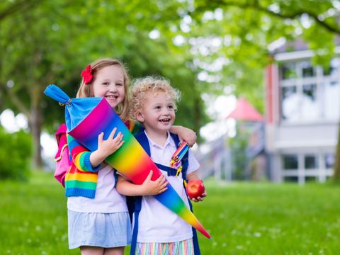 Zwei lachende Kinder mit einer regenbogenfarbenen Schultüte im Arm auf einer Wiese