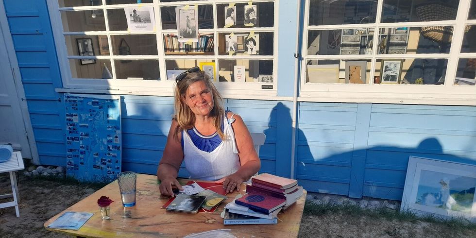 Das Bild zeigt die Autorin Ute Fritsch vor ihrem Haus auf Hiddensee. Sie sitzt umringt von ihren Kunstwerken und Büchern. 