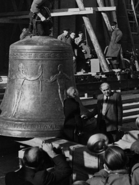 Enlarge photo: Several photographers take photos of a bell stored on a wooden platform and a man standing next to it. 