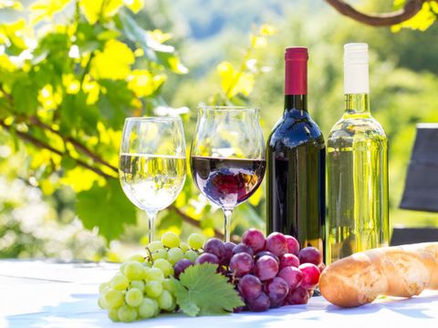 Ein Tisch im freien auf dem 2 verschiedene Sorten Wein in 2 Flaschen und in 2 Gläsern stehen, davor 2 Sorten Weintrauben und ein Baguette