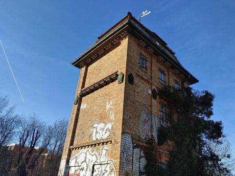 Ein schräg von unten vor blauem Himmel aufgenommenes, altes, hohes rechteckiges Gebäude aus Backsteinen mit Grafiti an den Wänden und mit einer Wetterfahne auf dem Dach.