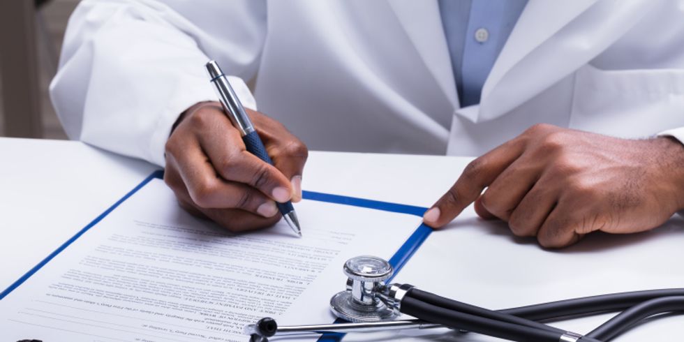 Ein Arzt sitzt an einem Tisch und schreibt mit einem Stift auf einem Blatt Papier in einem Klemmbrett. Daneben liegt ein Stethoskop.