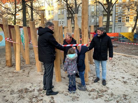 Bildvergrößerung: Zwei Männer und eine Frau stehen zusammen mit zwei Kindern auf einem Spielplatz mit aufgestellten Baumstämmen. Um die Stämme ist ein Absperrband, das von den Kindern durchgeschnitten wird.