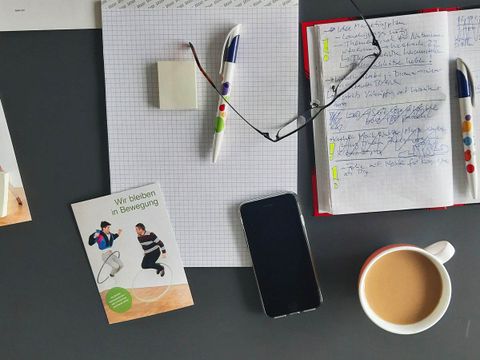 ein Schreibtisch von oben mit Schreibblock, Brille, Kaffeetasse, Handy, Stift und Postkarte mit Kampagnenmotiv