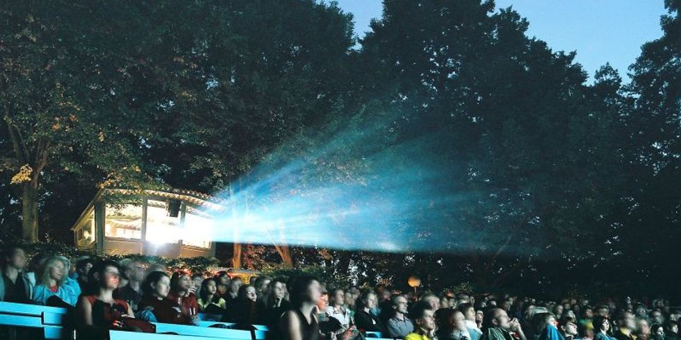 Freiluftkino Hasenheide bei Nacht mit Bäumen im Hintergrund. Der Lichtstrahl des Filmprojektors leuchtet über die Köpfe der Besucher und Besucherinnen