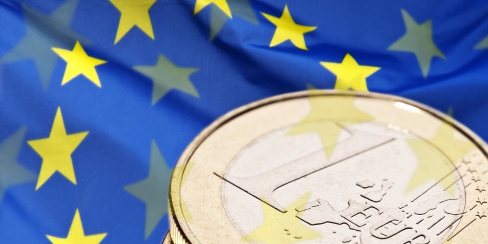 Übereinander liegende Ein-Euro-Münzen. Im Hintergrund sieht man die Europaflagge.