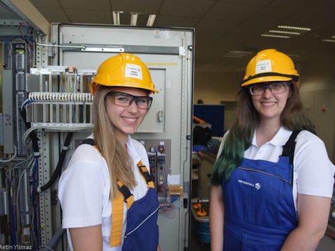 Bildvergrößerung: 2 Berliner Girls’Day Botschafterinnen stehen lächelnd in Arbeitslatzhose und Helm auf dem Kopf vor einem geöffneten Stromkasten
