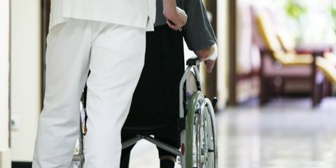 Auf der Pflegestation schiebt ein Pfleger einen Menschen mit Behinderung im Rollstuhl.