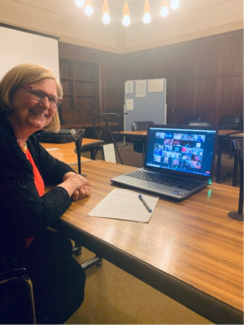 Bildvergrößerung: Eine Frau sitzt lächelnd an einem Schreibtisch. Auf dem Schreibtisch steht ein Laptop, auf dem eine Videokonferenz zu sehen ist.