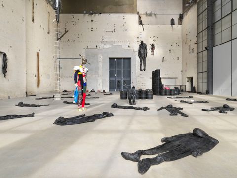 Kunstausstellung. In einem weißen Raum liegen mehrere schwarze Overalls auf dem Boden verteilt. In der Bildmitte steht eine Schaufesnterpuppe ohne Kopf, die bunte Kleidung trägt