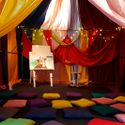 Bildvergrößerung: Blick in ein von einer Lichterkette beleuchtetes Zelt aus bunten Stoffbahnen, im Inneren steht eine Staffelei mit einem bunten Bild, Kissen liegen auf dem mit Teppich ausgelegten Boden