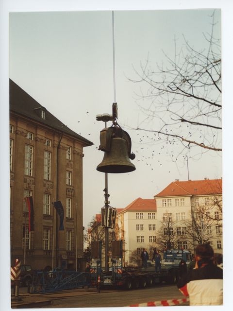 Enlarge photo: A large bell is hanging on a steel cable in front of a large building.