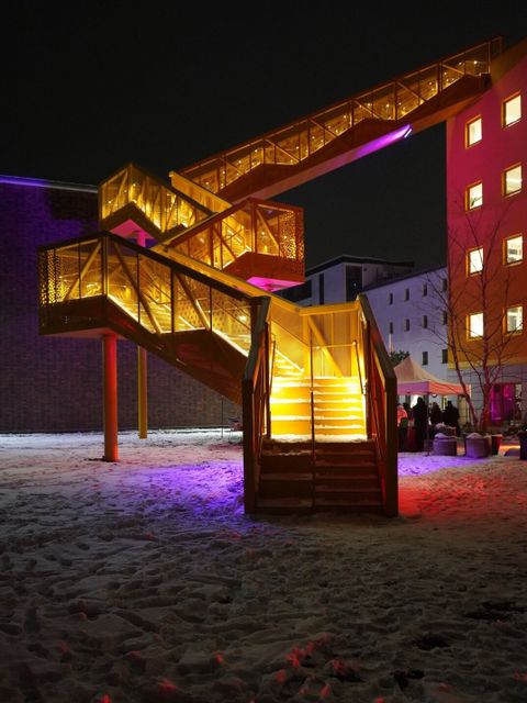 Bildvergrößerung: Nachts führt eine gelb beleuchtete Zick-Zack-Treppe von einem Platz hoch in ein Gebäude.