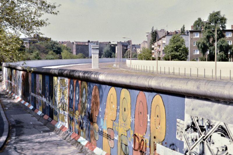 Bogen des Mauerstreifens entlang des Bethaniendamms, aufgenommen aus erhöhter Position mit Blick in den Grenzstreifen auf einen Wachturm. Im Vordergrund sind Mauerelemente mit den charakteristischen "Köpfen" von Thierry Noir zu sehen.