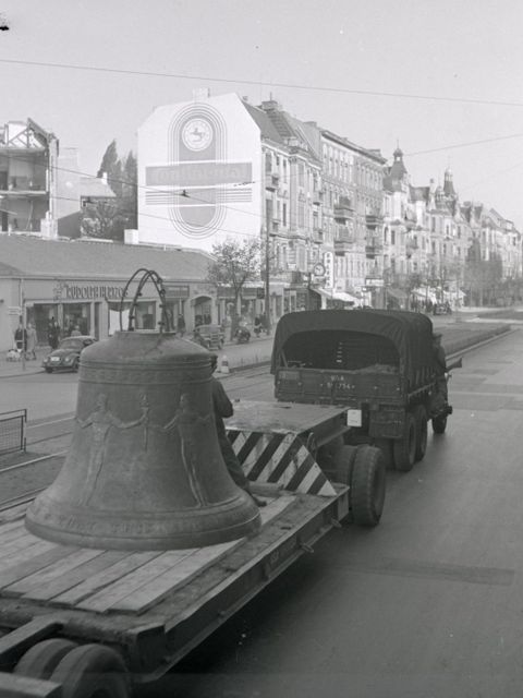 Enlarge photo: A large cast iron bell on a flat trailer is being pulled through the city by a car.