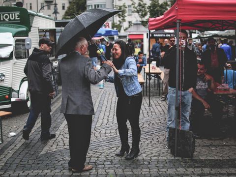 Auf der rechten Bildseite mehrere Bierbankgarnituren an denen Personen sitzen. Auf der linken Bildseite eine Frau mit einer hellen Jeansjacke und einem schwarzen T-Shirt und einer schwarzen Hose, die gerade einen älteren Herren mit einem Regenschirm anspricht. 