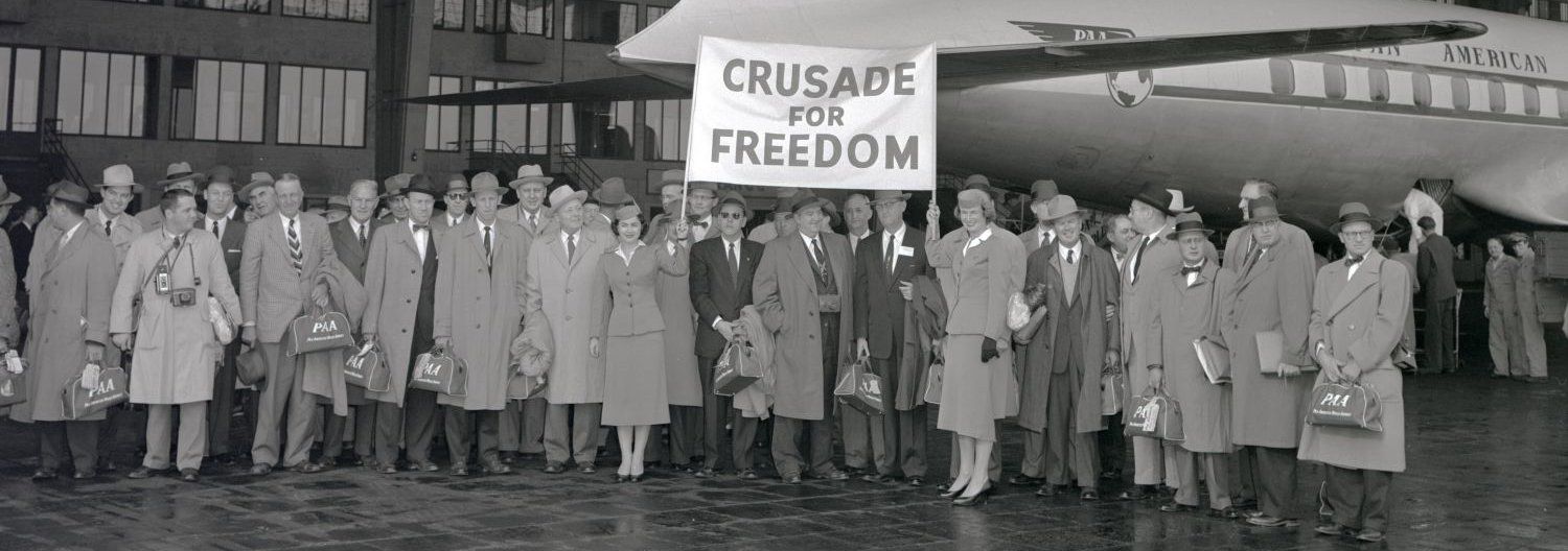 Enlarge photo: A large group of people in an aircraft hangar, with an airplane in the background. A sign is held up with the inscription “Crusade for Freedom”. 