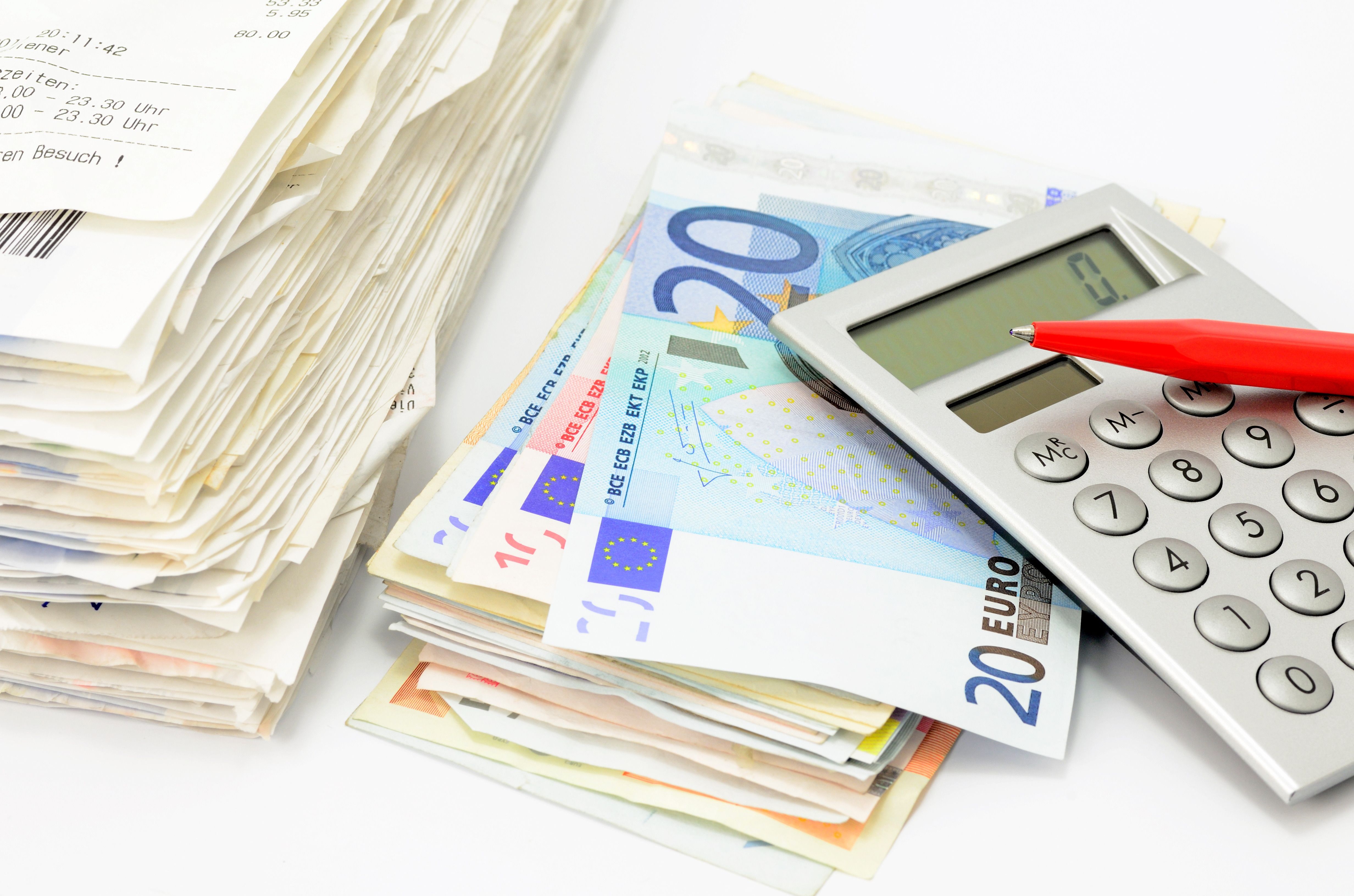 Das Bild zeigt einen Rechnungs- und einen Geldstapel mit Taschenrechner und Stift