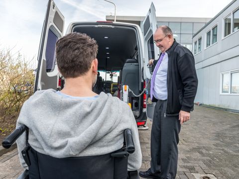 Ein Mann hilft einem männlichen Rollstuhlfahrer über eine an Hebebühne an Bord eines Kleinbusses zu kommen
