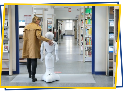 Eine Frau und ein Roboter stehen vor den Gängen der Spandauer Bibliothek
