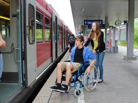 Mädchen schiebt Teenager im Rollstuhl Richtung wartendem Zug am Bahnsteig