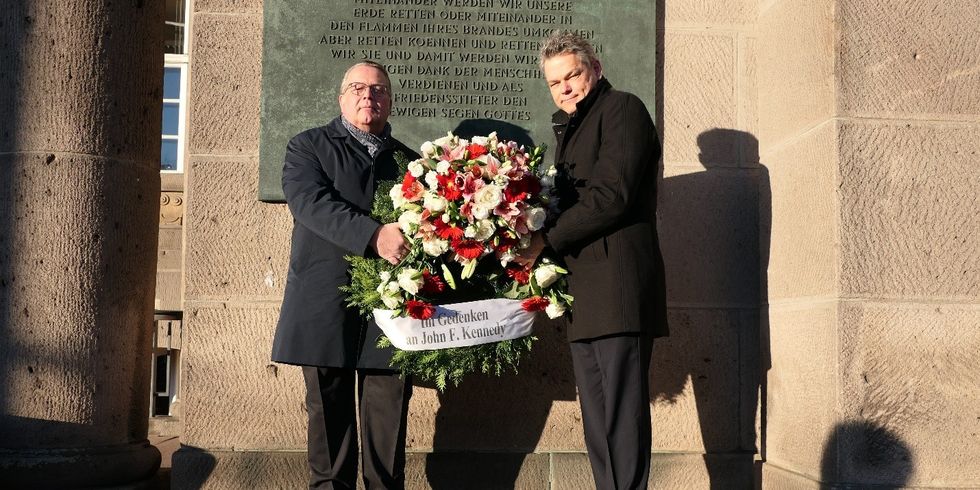 Zwei Männer stehen vor einer Gedenktafel und halten einen großen Kranz mit einem Band mit der Aufschrift "Im Gedenken an John-F.-Kennedy".