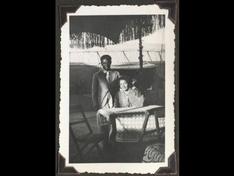 Bildvergrößerung: Altes Schwarz-Weiß-Foto. In einem Garten sitzt eine Frau an einem Tisch und ein Mann steht neben ihr.