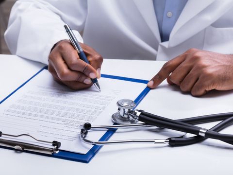 Ein Arzt sitzt an einem Tisch und schreibt mit einem Stift auf einem Blatt Papier in einem Klemmbrett. Daneben liegt ein Stethoskop.