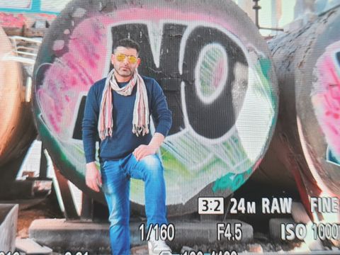 Bildvergrößerung: Ein Mann mit Sonnenbrille und dünnem Schal steht lässig vor einem Grafitti. Rechts unten sieht man die Kameraeinstellungen mit denen das Bild gemacht wurde.