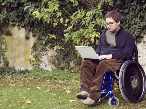 Ein junger Rollstuhlfahrer sitzt mit Laptop auf dem Schoß in der freien Natur