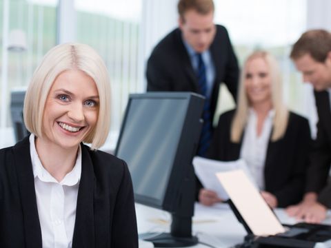 Lächelnde Geschäftsfrau am Schreibtisch mit ihren Kollegen im Hintergrund 
