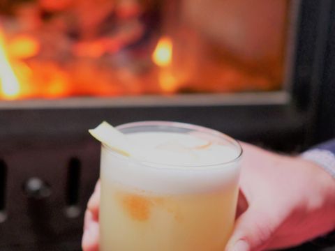 Eine Hand hält ein gefülltes Cocktailglas. Die Flüssigkeit im Glas ist hellgelb und schaumig. Im Hintegrund brennt ein Kamin