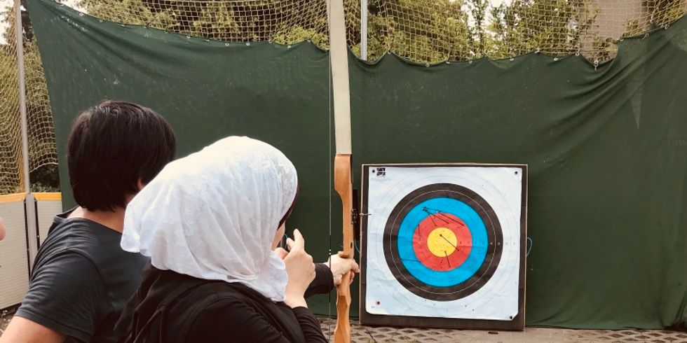 Eine Frau mit Kopftuch spannt Pfeil und Bogen und zielt auf eine bunte Zielscheibe
